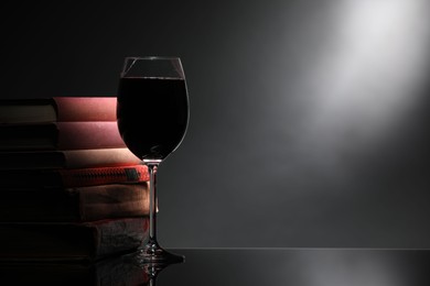 Photo of Tasty wine in glass and stack of books on mirror table against dark background. Space for text