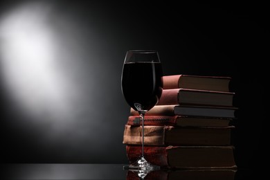 Photo of Tasty wine in glass and stack of books on mirror table against dark background. Space for text