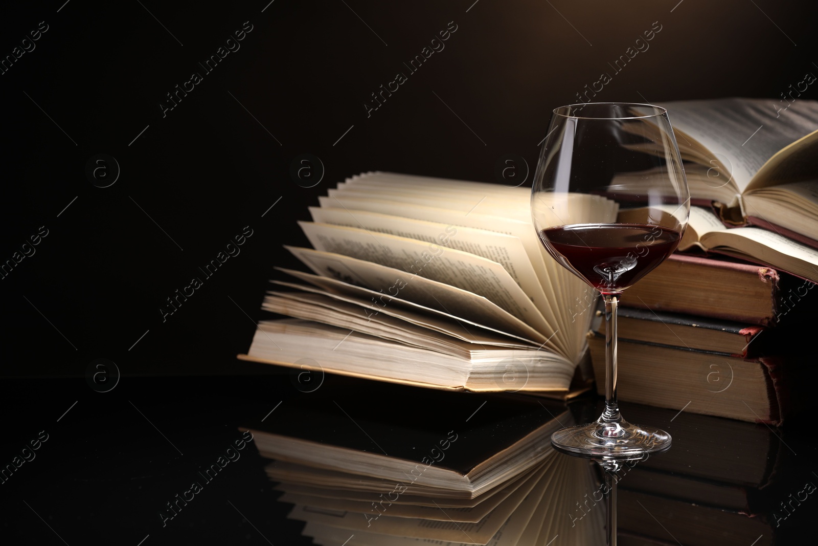 Red wine in glass and stack of books on mirror table against dark background, closeup Photo of Red wine in glass and stack of books on mirror table against dark background, closeup