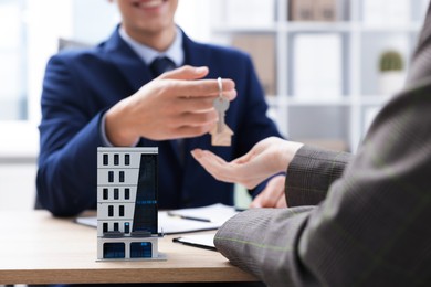 Photo of Real estate agent giving key to client at desk in office, focus on house model
