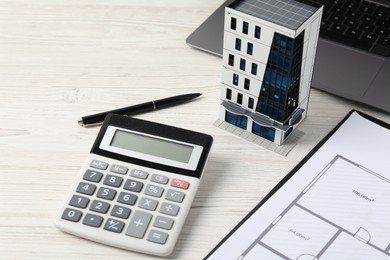 Photo of Real estate. Apartment building model, floor plan, calculator and laptop on white wooden table, closeup