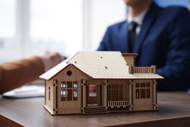 Photo of Real estate agent and client shaking hands at desk in office, focus on house model