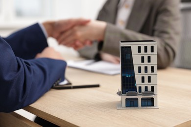 Photo of Real estate agent and client shaking hands at desk in office, focus on house model
