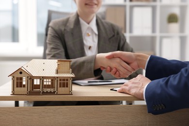 Photo of Real estate agent and client shaking hands at desk in office, focus on house model