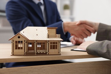Photo of Real estate agent and client shaking hands at desk in office, focus on house model