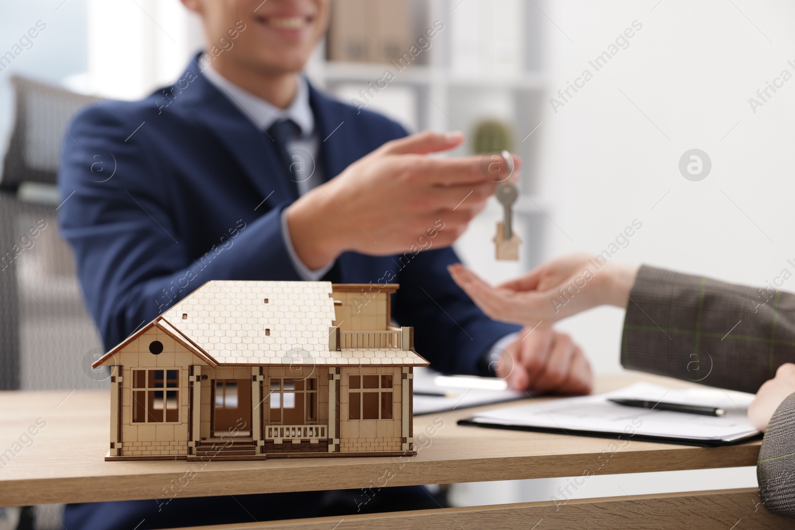 Real estate agent giving key to client at desk in office, focus on house model Photo of Real estate agent giving key to client at desk in office, focus on house model