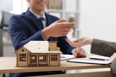 Photo of Real estate agent giving key to client at desk in office, focus on house model