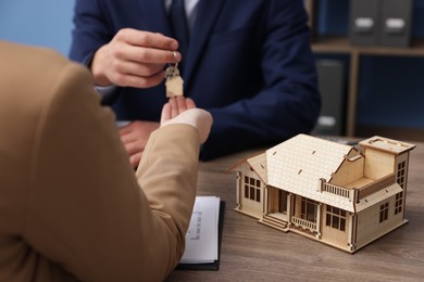 Photo of Real estate agent giving key to client at desk in office, focus on house model