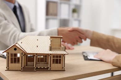 Photo of Real estate agent and client shaking hands at desk in office, focus on house model