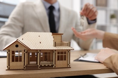 Photo of Real estate agent giving key to client at desk in office, focus on house model