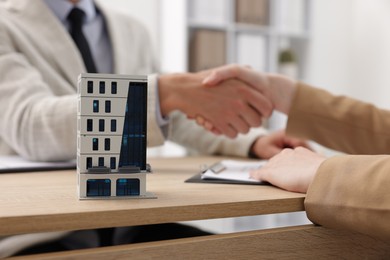 Photo of Real estate agent and client shaking hands at desk in office, focus on house model