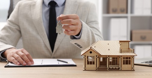 Photo of Real estate agent holding key at desk in office, focus on house model
