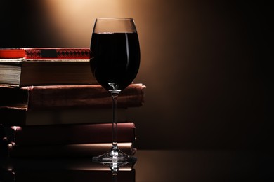 Photo of Tasty wine in glass and stack of books on mirror table against dark background, closeup. Space for text