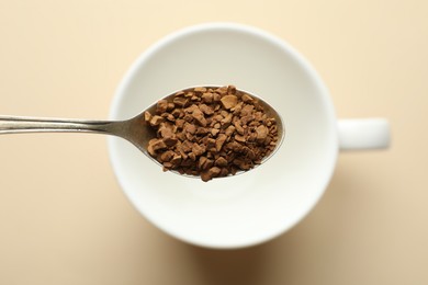 Photo of Spoon with instant coffee above cup on beige background, top view
