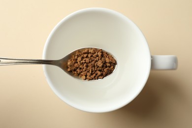 Photo of Spoon with instant coffee above cup on beige background, top view