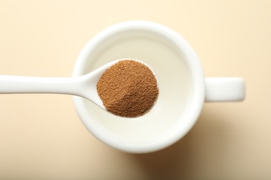 Photo of Spoon with instant coffee above cup on beige background, top view