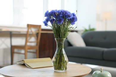 Photo of Beautiful blue cornflowers in vase and book on wooden table at home