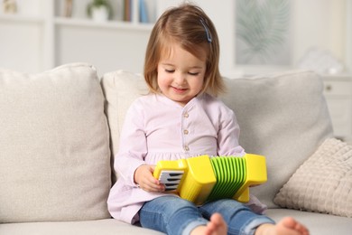 Photo of Cute little girl playing toy accordion on sofa at home