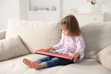 Photo of Cute little girl playing toy electric piano on sofa at home