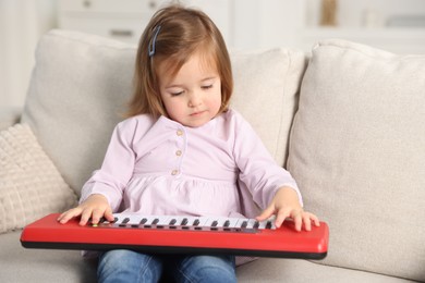 Photo of Cute little girl playing toy electric piano on sofa at home