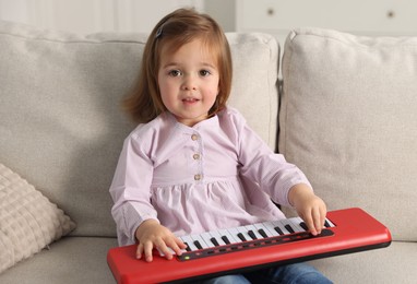 Photo of Cute little girl playing toy electric piano on sofa at home