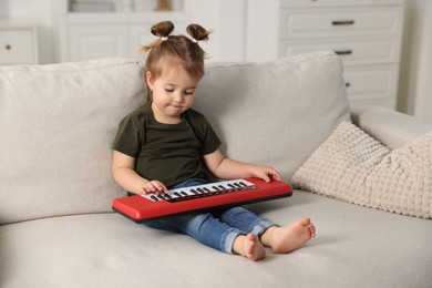 Photo of Cute little girl playing toy electric piano on sofa at home
