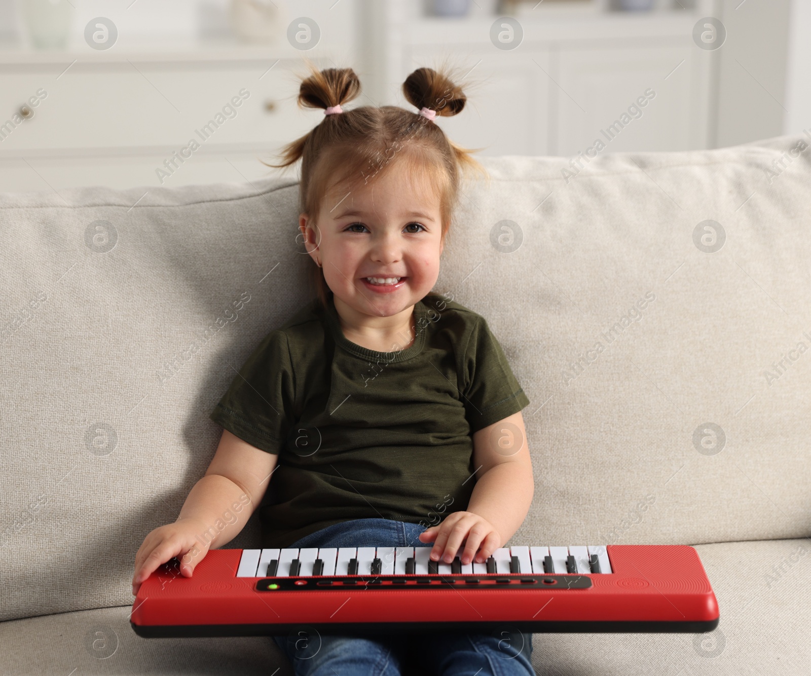 Photo of Cute little girl playing toy electric piano on sofa at home