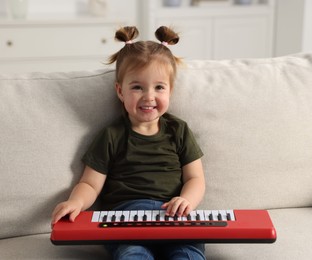 Photo of Cute little girl playing toy electric piano on sofa at home