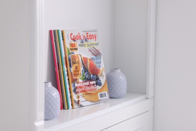 Photo of Many different trendy magazines and vases in white shelving unit indoors