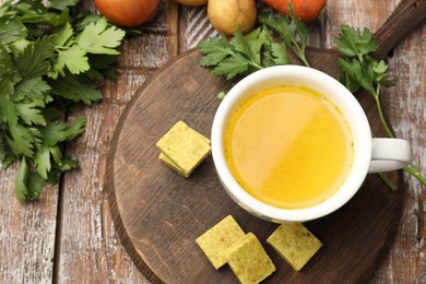 Photo of Aromatic bouillon cubes, broth, parsley and vegetables on wooden table, flat lay