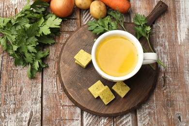 Photo of Aromatic bouillon cubes, broth, parsley and vegetables on wooden table, flat lay