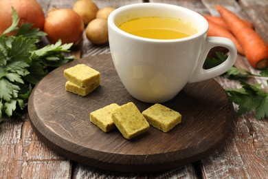 Photo of Aromatic bouillon cubes, broth, parsley and vegetables on wooden table, closeup
