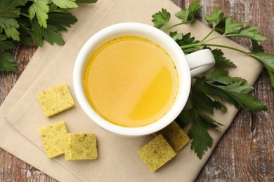 Photo of Aromatic bouillon cubes, broth and parsley on wooden table, flat lay