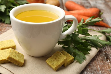 Photo of Aromatic bouillon cubes, broth, parsley and vegetables on wooden table, closeup