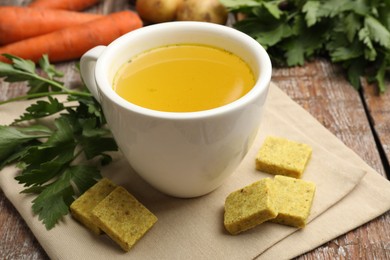 Photo of Aromatic bouillon cubes, broth, parsley and vegetables on wooden table, closeup