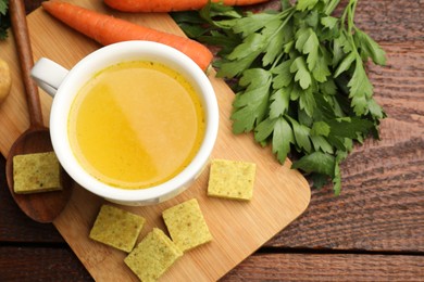 Photo of Aromatic bouillon cubes, broth, parsley and vegetables on wooden table, flat lay