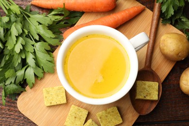 Photo of Aromatic bouillon cubes, broth, parsley and vegetables on wooden table, flat lay