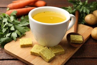 Photo of Aromatic bouillon cubes, broth, parsley and vegetables on wooden table, closeup