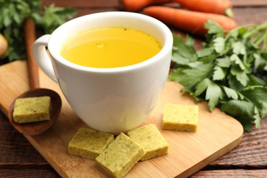 Photo of Aromatic bouillon cubes, broth, parsley and vegetables on wooden table, closeup