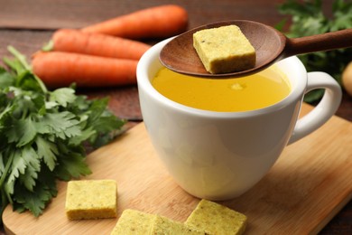 Photo of Putting aromatic bouillon cube into broth at wooden table, closeup