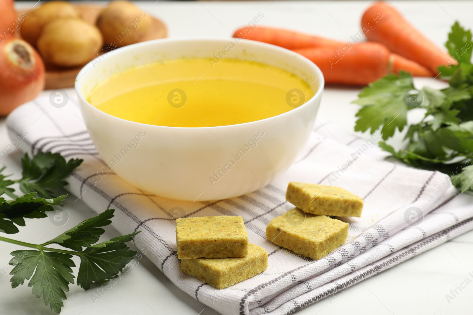 Photo of Aromatic bouillon cubes, broth, parsley and vegetables on white table, closeup