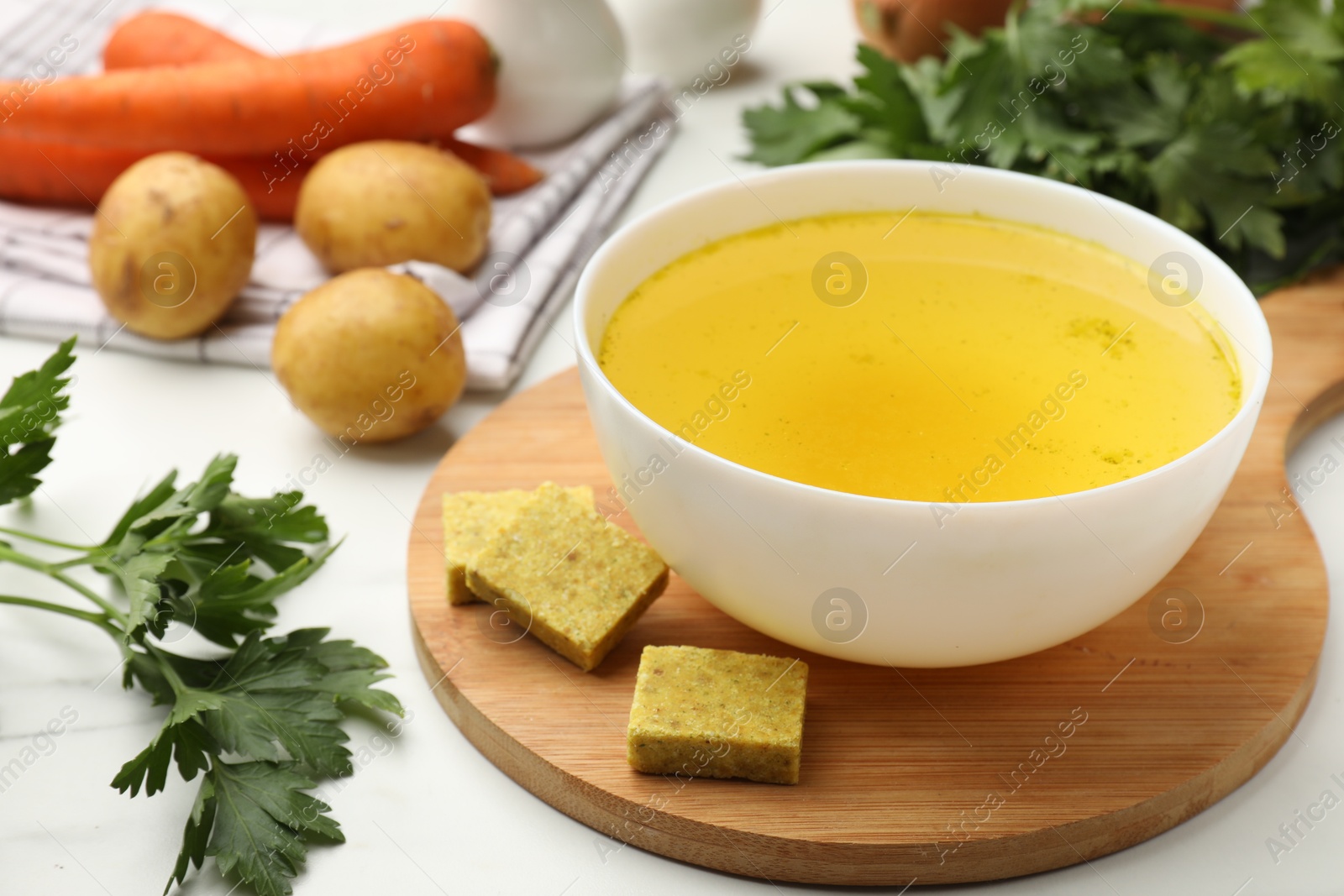 Photo of Aromatic bouillon cubes, broth, parsley and vegetables on white marble table, closeup