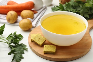 Photo of Aromatic bouillon cubes, broth, parsley and vegetables on white marble table, closeup