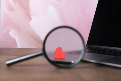 Photo of Decorative heart, magnifying glass and laptop on wooden table, closeup. Online dating site