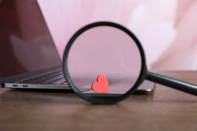 Photo of Decorative heart, magnifying glass and laptop on wooden table, closeup. Online dating site