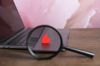 Photo of Decorative heart, magnifying glass and laptop on wooden table, closeup. Online dating site