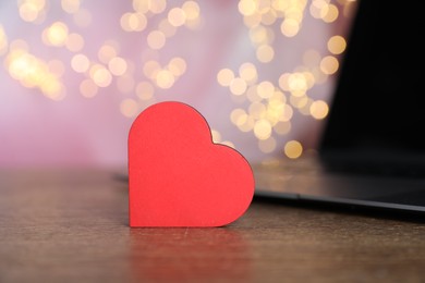 Photo of Online dating site. Decorative heart and laptop on wooden table against blurred lights, closeup. Bokeh effect
