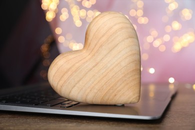 Photo of Online dating site. Wooden heart and laptop on table against blurred lights, closeup. Bokeh effect