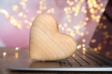 Photo of Online dating site. Wooden heart and laptop on table against blurred lights, closeup. Bokeh effect