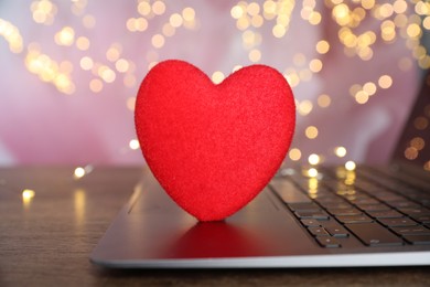 Photo of Online dating site. Decorative heart and laptop on wooden table against blurred lights, closeup. Bokeh effect